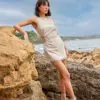 This dress is a sleeveless, natural beige mini-dress made from a lightweight fabric. It features a gathered, ruched detail at the side with an open crochet accent, adding a unique and stylish texture. The bottom hem has a delicate fringe trim, giving the dress a relaxed, bohemian vibe. The simple round neckline and fitted silhouette highlight its minimalistic yet elegant design, perfect for casual or semi-formal summer wear.