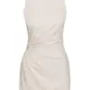 This dress is a sleeveless, natural beige mini-dress made from a lightweight fabric. It features a gathered, ruched detail at the side with an open crochet accent, adding a unique and stylish texture. The bottom hem has a delicate fringe trim, giving the dress a relaxed, bohemian vibe. The simple round neckline and fitted silhouette highlight its minimalistic yet elegant design, perfect for casual or semi-formal summer wear.