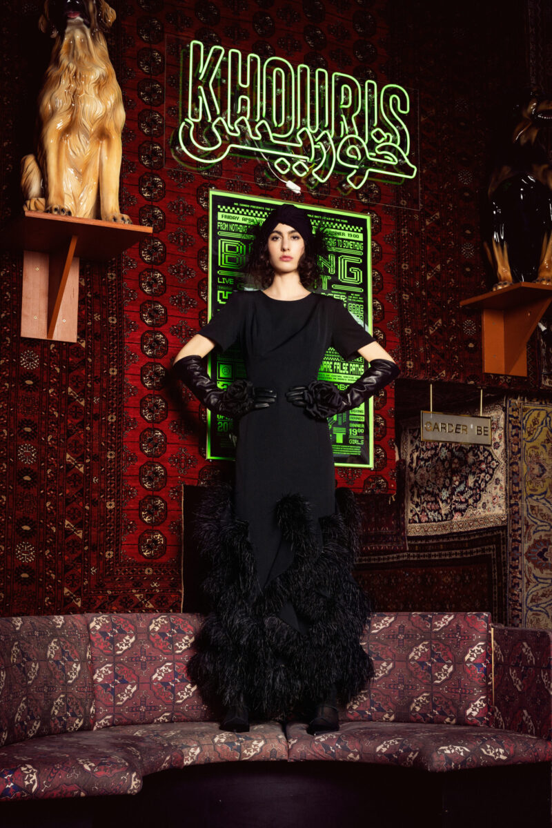 A woman in a black dress and gloves stands in a room with ornate rugs and a neon sign reading "KHOJURIS.