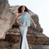 Model wearing the Nana Gotti Selin Crepe-Back Satin One-Shoulder Fringe Maxi Dress in powder blue, featuring cascading asymmetric fringe, a fitted silhouette, and a flared maxi hem, photographed against a natural rocky backdrop.