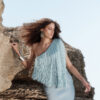 Model wearing the Nana Gotti Selin Crepe-Back Satin One-Shoulder Fringe Maxi Dress in powder blue, featuring dramatic cascading fringe, an asymmetric neckline, and a fitted satin silhouette, photographed against a natural rocky backdrop.