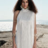 Close-up front view of the Nana Gotti Ay Viscose Satin Fringe Maxi Dress in ivory, featuring a high halter neckline, delicate cascading fringe, and a sleek satin silhouette, modeled against a sunlit coastal backdrop.