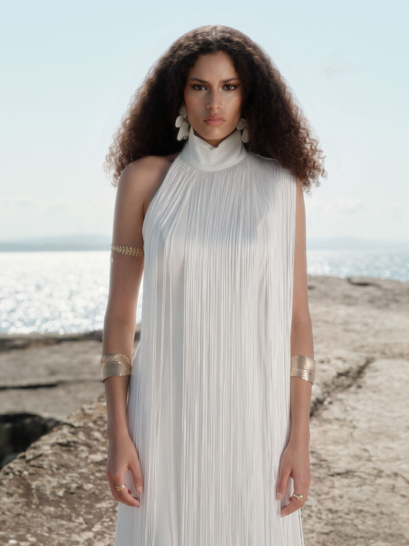 Close-up front view of the Nana Gotti Ay Viscose Satin Fringe Maxi Dress in ivory, featuring a high halter neckline, delicate cascading fringe, and a sleek satin silhouette, modeled against a sunlit coastal backdrop.