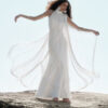 Model wearing the Nana Gotti Ay Viscose Satin Fringe Maxi Dress in ivory, featuring a high halter neckline, floor-length satin silhouette, and dramatic flowing fringe extending outward in motion against a sunlit rocky coastal backdrop.