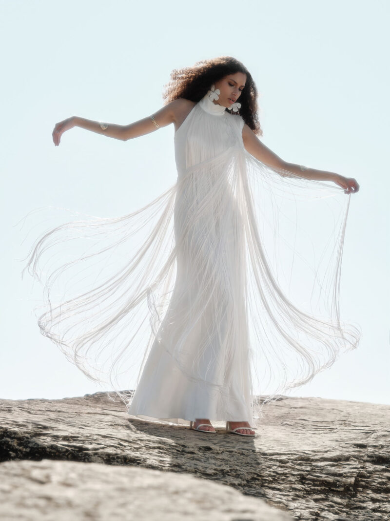 Model wearing the Nana Gotti Ay Viscose Satin Fringe Maxi Dress in ivory, featuring a high halter neckline, floor-length satin silhouette, and dramatic flowing fringe extending outward in motion against a sunlit rocky coastal backdrop.