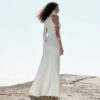 Back and side view of the Nana Gotti Ay Viscose Satin Fringe Maxi Dress in ivory, featuring a high halter neckline, sleek fitted waist, flowing floor-length satin skirt, and delicate fringe cascading down the back, photographed on a sunlit rocky coastline.