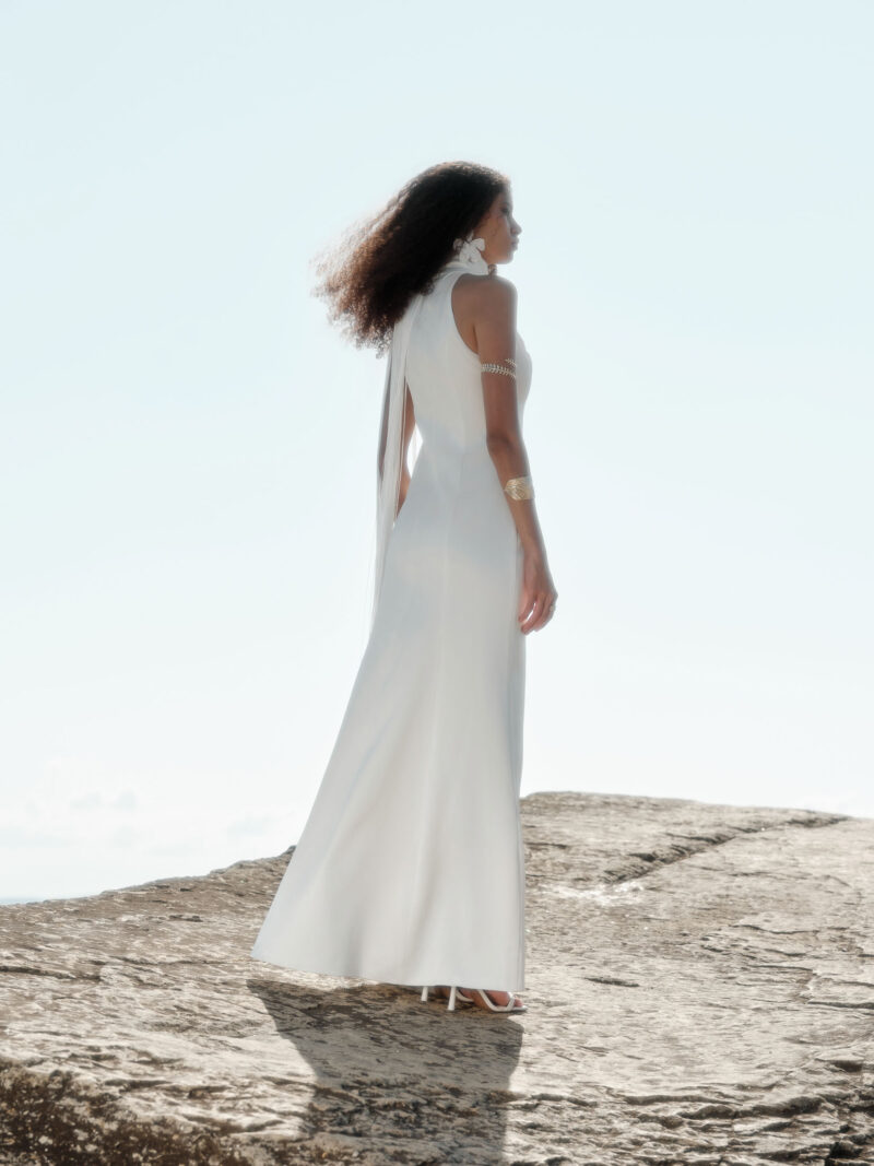 Back and side view of the Nana Gotti Ay Viscose Satin Fringe Maxi Dress in ivory, featuring a high halter neckline, sleek fitted waist, flowing floor-length satin skirt, and delicate fringe cascading down the back, photographed on a sunlit rocky coastline.