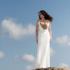 Model wearing the Nana Gotti Aurore Off-White Viscose Crepe Chain-Drape Maxi Dress, featuring delicate spaghetti straps, a draped gold-tone chain neckline, and a fitted floor-length silhouette against a coastal backdrop.