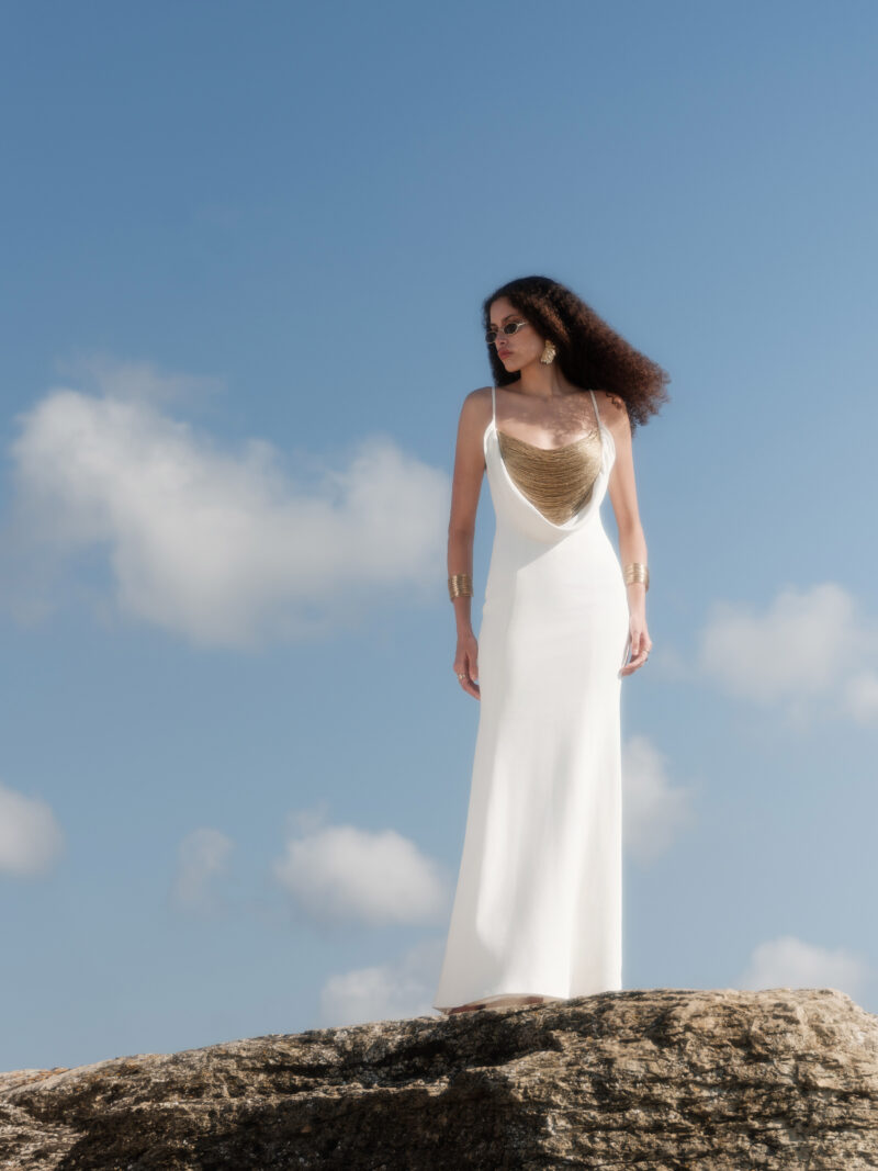 Model wearing the Nana Gotti Aurore Off-White Viscose Crepe Chain-Drape Maxi Dress, featuring delicate spaghetti straps, a draped gold-tone chain neckline, and a fitted floor-length silhouette against a coastal backdrop.
