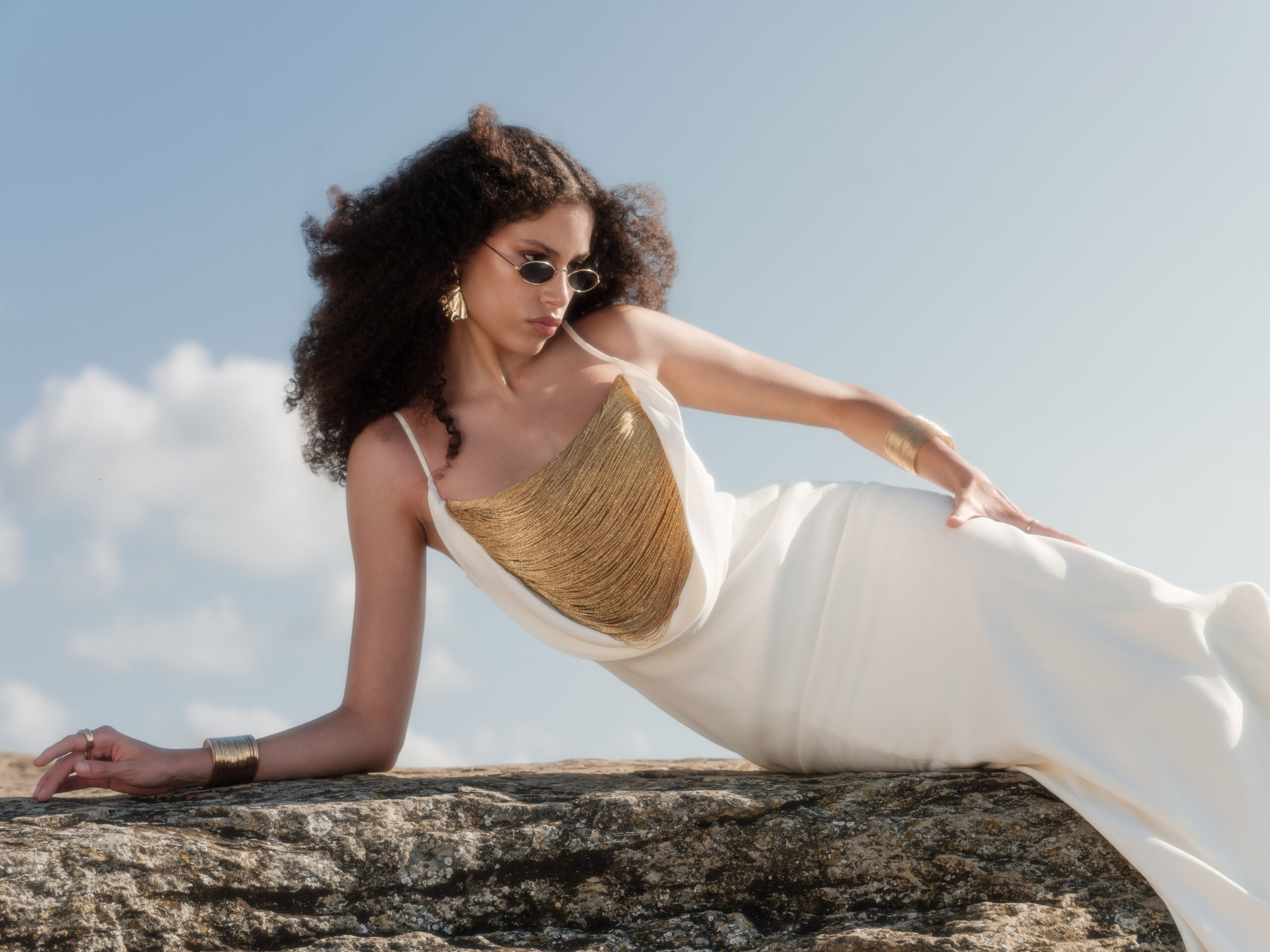 Model wearing the Nana Gotti Aurore Off-White Maxi Dress with a draped gold fringe neckline and flowing silhouette, reclining on rocks under a blue sky.