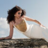 Model wearing the Nana Gotti Aurore Off-White Viscose Crepe Chain-Drape Maxi Dress with cascading gold-tone chain detailing and delicate spaghetti straps, reclining on coastal rocks.