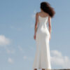 Back view of model wearing the Nana Gotti Aurore Off-White Viscose Crepe Chain-Drape Maxi Dress, featuring delicate spaghetti straps, a fitted silhouette, and a softly flared floor-length skirt against a coastal backdrop