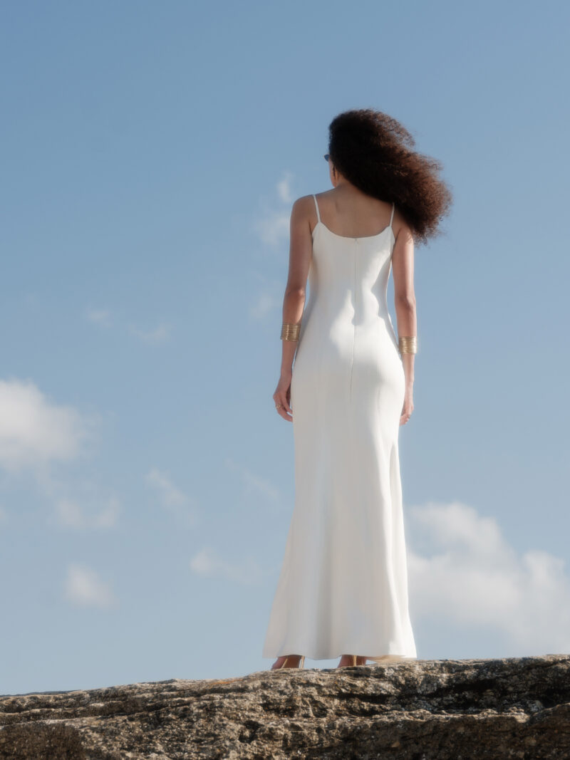 Back view of model wearing the Nana Gotti Aurore Off-White Viscose Crepe Chain-Drape Maxi Dress, featuring delicate spaghetti straps, a fitted silhouette, and a softly flared floor-length skirt against a coastal backdrop
