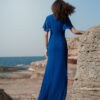 Back view of a model wearing the Nana Gotti Azura cobalt blue maxi gown with chiffon flutter sleeves and a flowing floor-length silhouette by the sea.