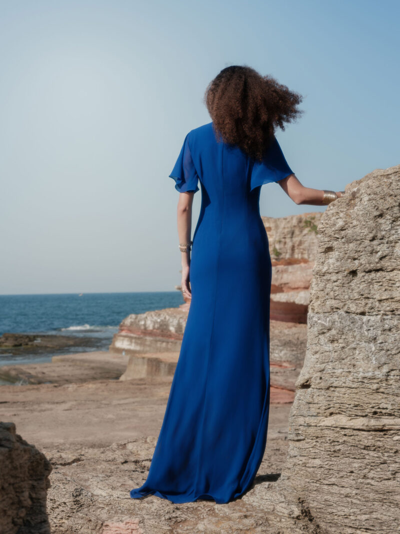 Back view of a model wearing the Nana Gotti Azura cobalt blue maxi gown with chiffon flutter sleeves and a flowing floor-length silhouette by the sea.