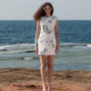 Full-length front view of a model wearing the Nana Gotti Narin ivory and blue floral lace mini dress with white heels on a rocky Mediterranean coastline.