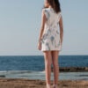 Model showcasing the back of the Nana Gotti Narin floral lace mini dress in ivory with blue İznik-inspired motifs against a coastal backdrop.