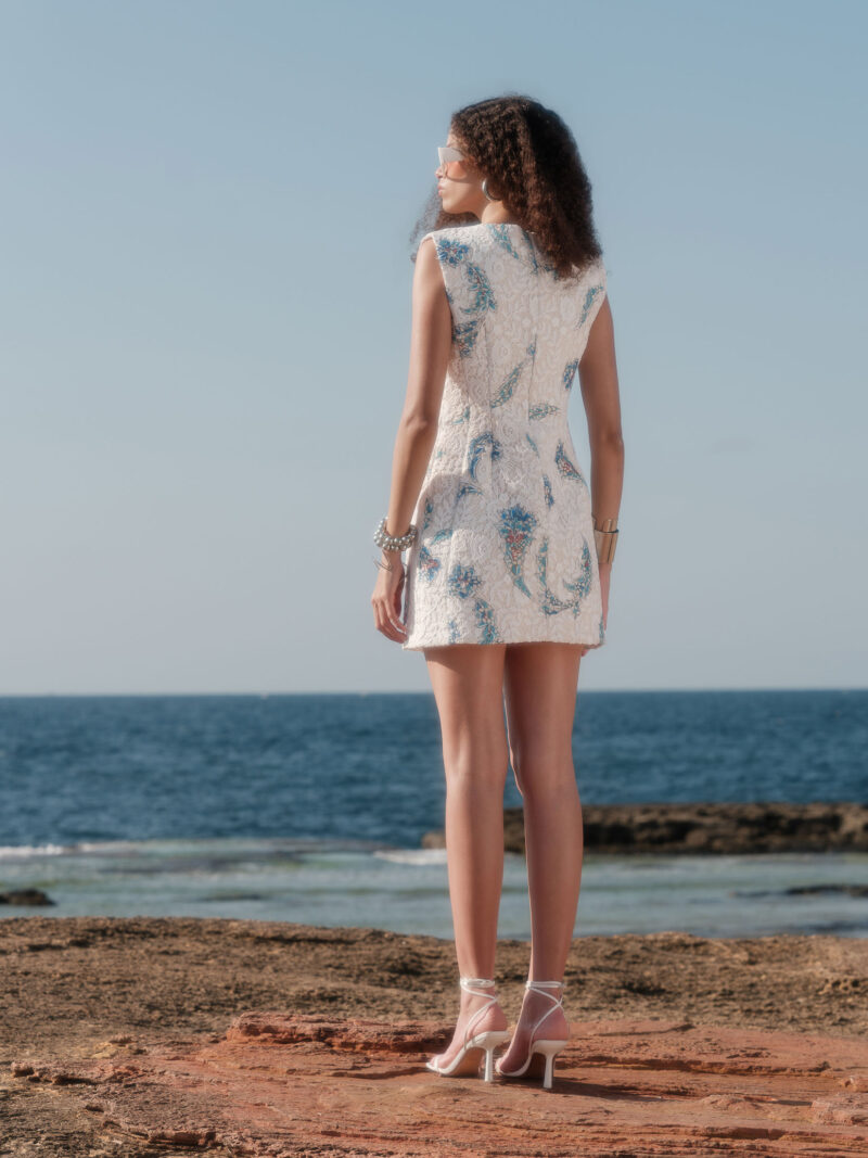 Model showcasing the back of the Nana Gotti Narin floral lace mini dress in ivory with blue İznik-inspired motifs against a coastal backdrop.