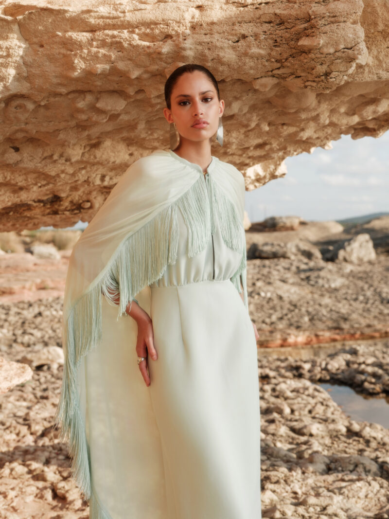 Model wearing the Nana Gotti Nerina Mint Green Fringe Cape Maxi Dress with cascading chiffon cape and fringe detailing, styled for luxury occasionwear against a natural rocky backdrop.