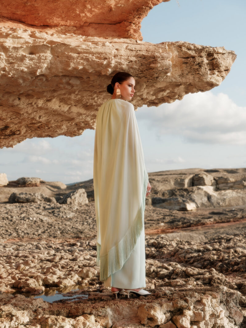 Model wearing the Nana Gotti Nerina Mint Green Fringe Cape Maxi Dress with cascading chiffon cape and fringe detailing, styled for luxury occasionwear against a natural rocky backdrop.