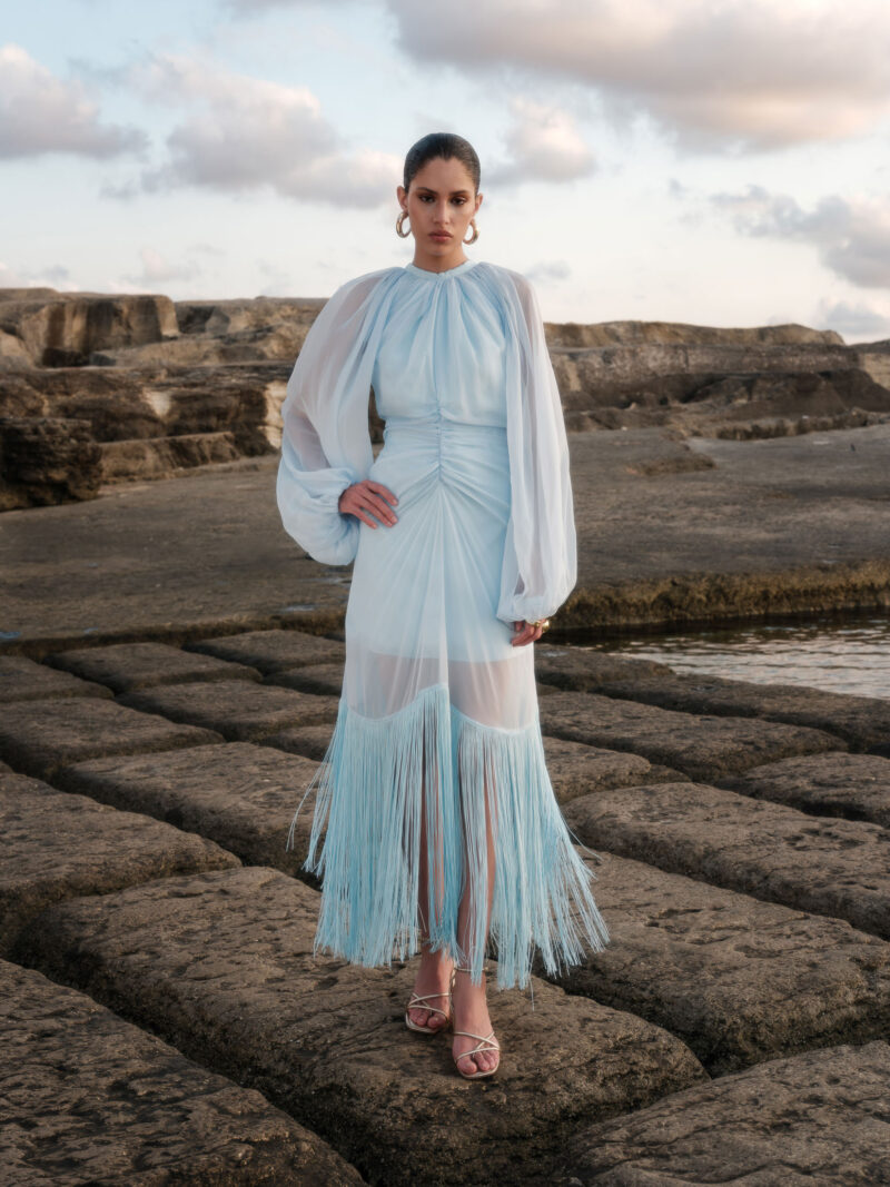 Ege Ruched Chiffon Fringe Midi Dress by Nana Gotti featuring a high neckline, voluminous sleeves, fitted waist, and cascading blue fringe hem.