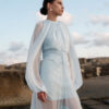 Nana Gotti Ege light blue chiffon fringe midi dress with sheer balloon sleeves, gathered neckline, and ruched waist, photographed by the sea.
