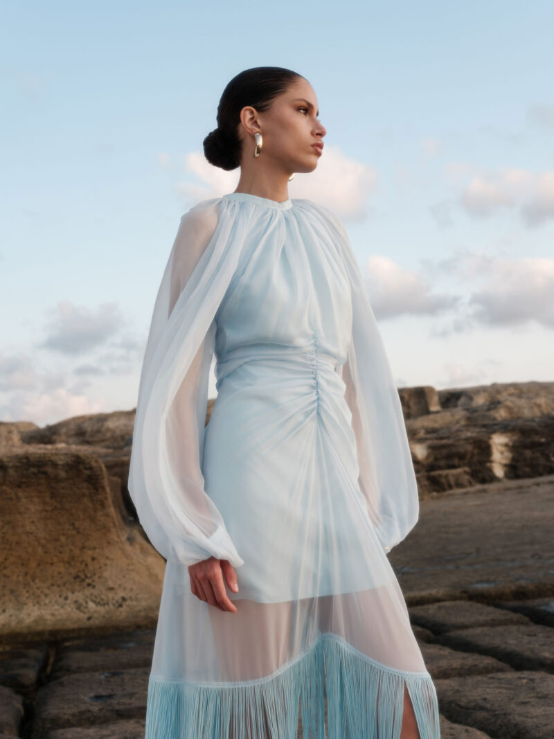 Nana Gotti Ege light blue chiffon fringe midi dress with sheer balloon sleeves, gathered neckline, and ruched waist, photographed by the sea.