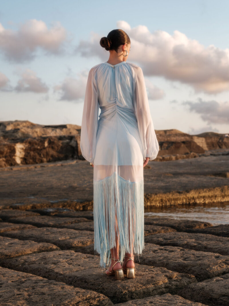 Back view of Nana Gotti Ege Fringe Midi Dress in pastel blue with sheer chiffon sleeves, ruched back, and flowing fringe detailing.