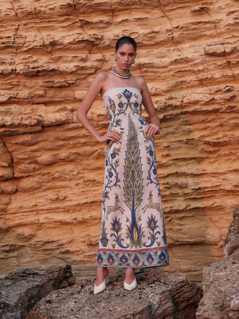 Model wearing the Nana Gotti Yara ivory printed linen strapless maxi dress with colorful Anatolian-inspired botanical motifs against a sandstone backdrop.